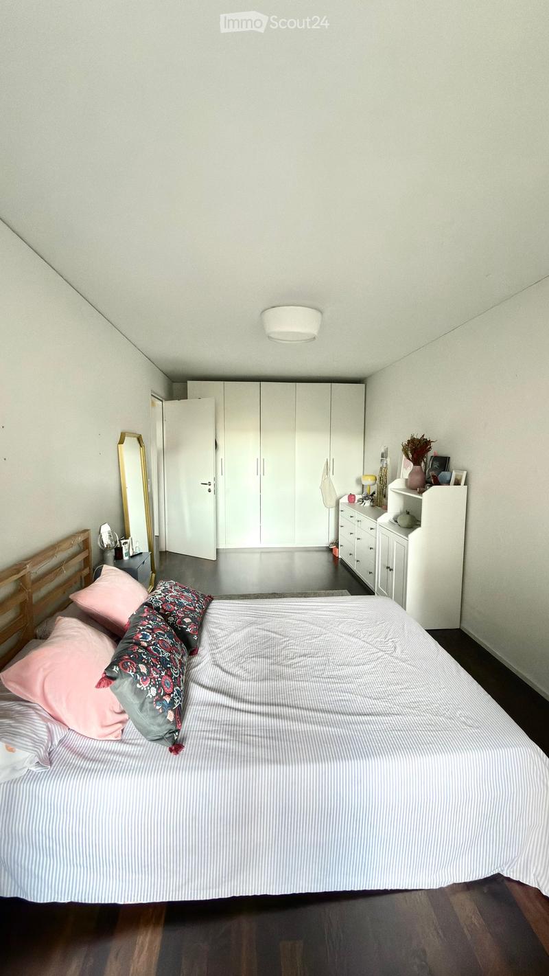 A modern bedroom with a large bed, striped bedding, pink and patterned pillows, a wooden headboard, white wardrobe, a gold-framed mirror, and a nightstand with a lamp and decorative items.