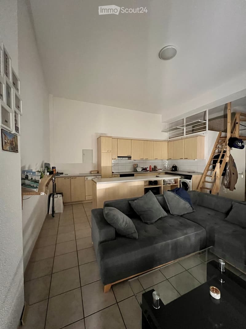 A modern, open-plan living and kitchen area with a grey sectional sofa, glass coffee table, kitchen cabinets, and a dishwasher.