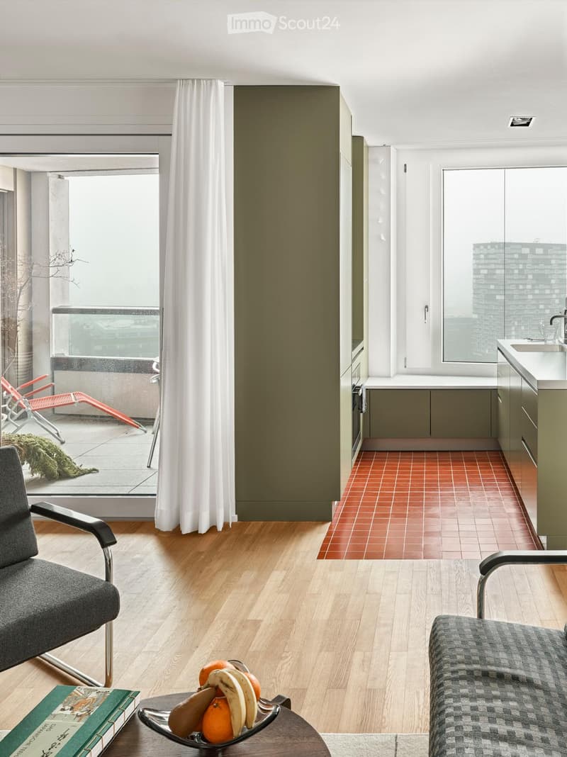 Modern living room with wooden flooring, grey sofa, armchair, coffee table with fruit bowl, and a view of a balcony with lounge chairs.
