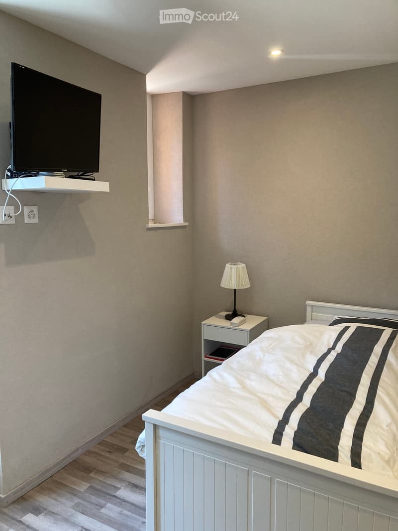 A modern bedroom with a white bed, nightstand, lamp, and a wall-mounted screen. The room features light-colored walls and wooden flooring.