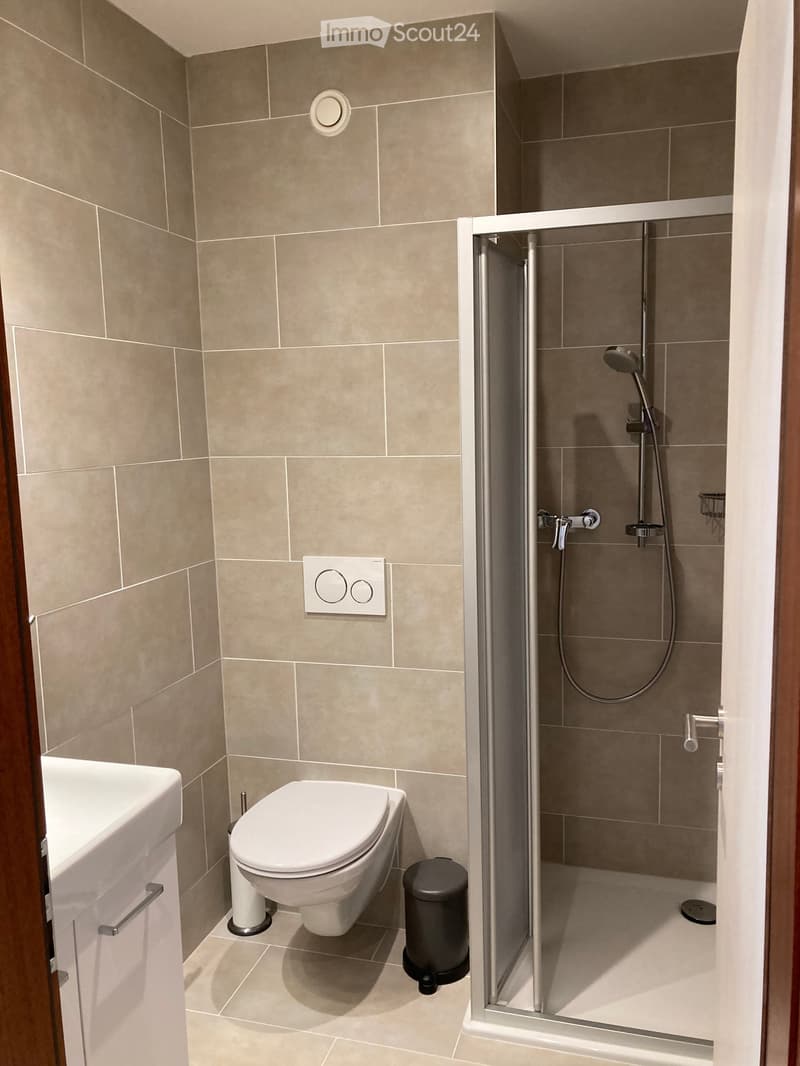 Modern bathroom with beige tiled walls and floor, featuring a toilet, sink, and a glass-enclosed shower with a handheld showerhead.