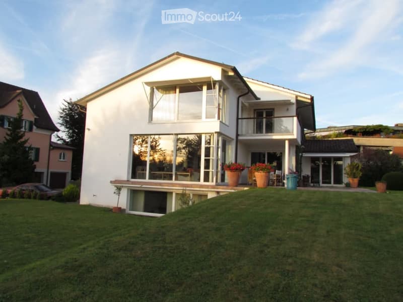 modern 2-story house, white exterior, large windows, balcony, raised ground floor, surrounded by a large lawn and greenery