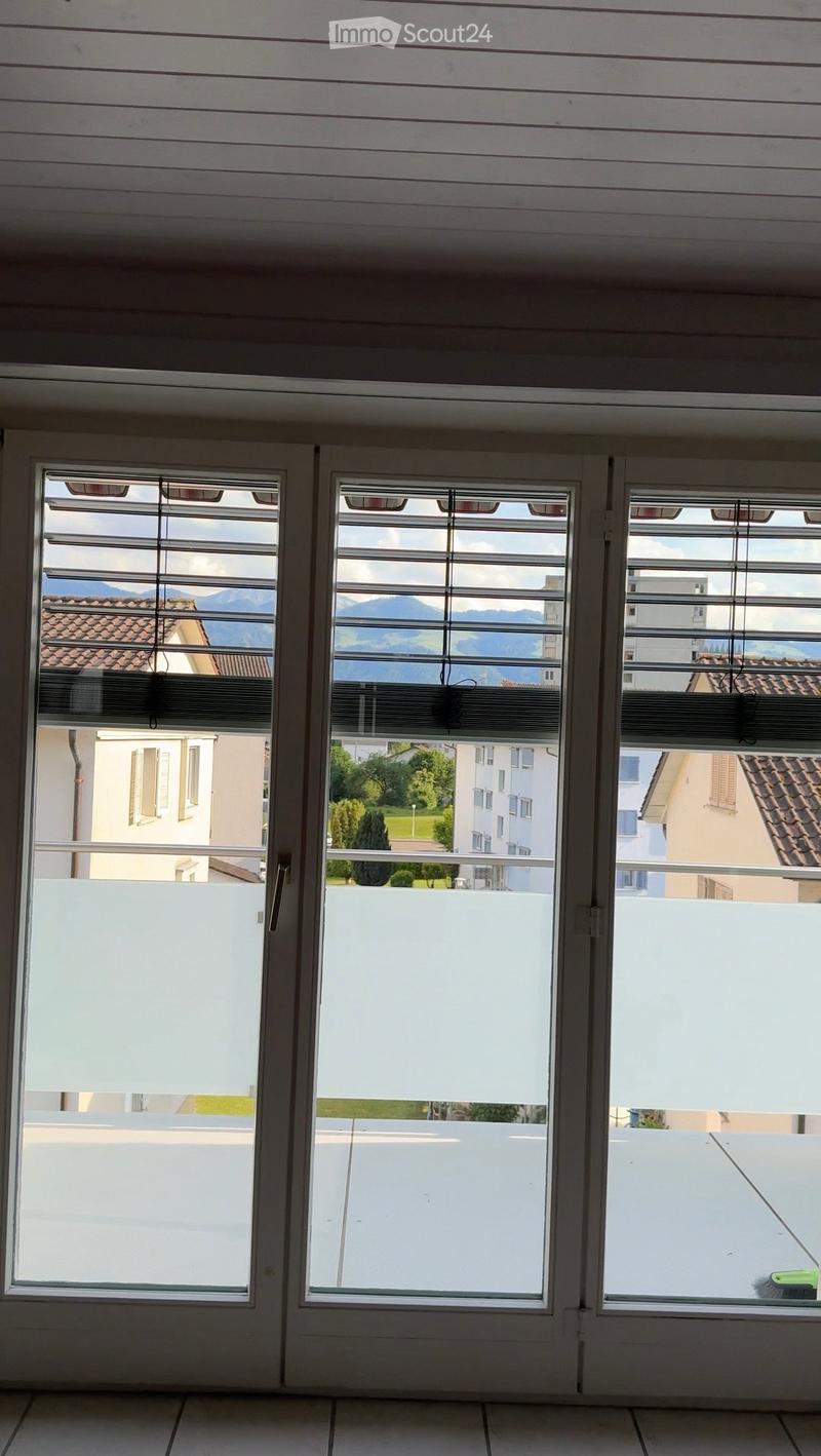 Empty room with large windows and blinds, overlooking a view of mountains and buildings. The floor is tiled, and the walls are painted white.