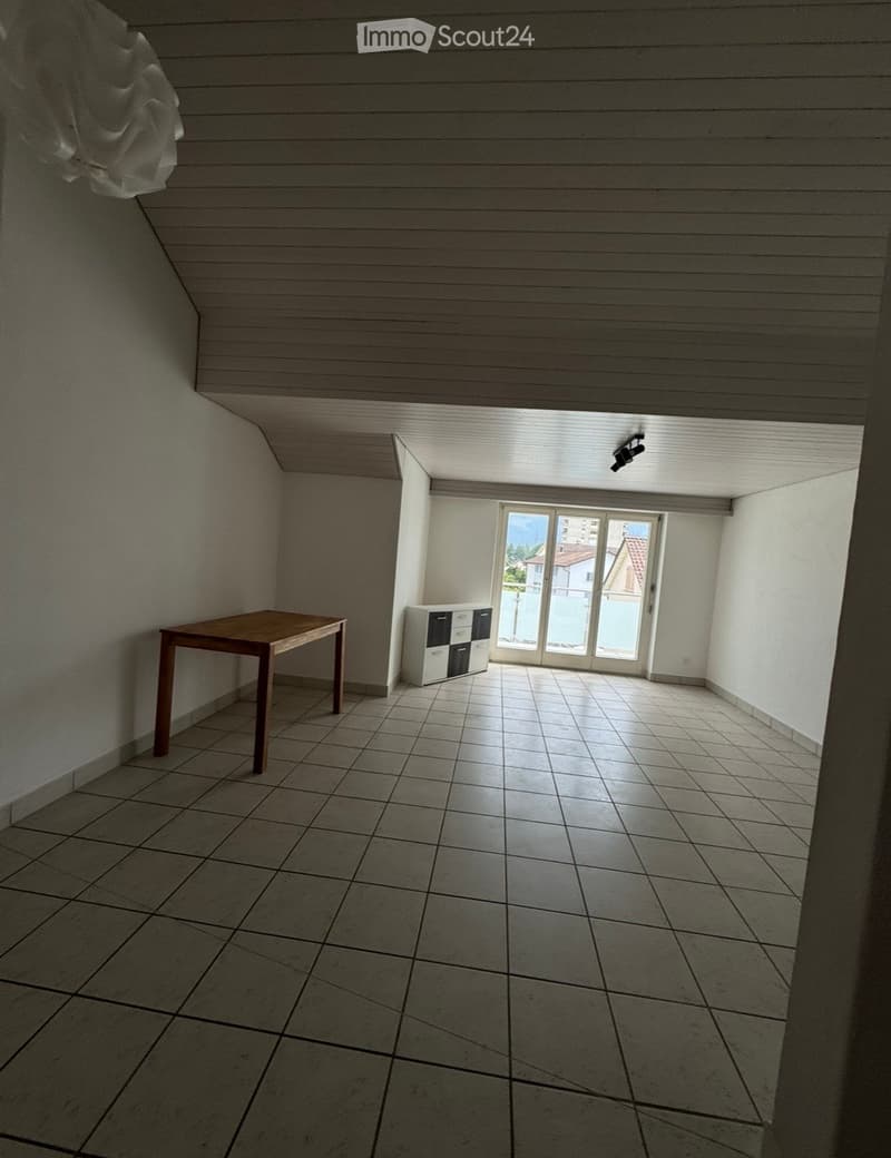 A small, unfurnished room with tiled flooring, a wooden table, and a small cabinet near a large window with a view of the outside.