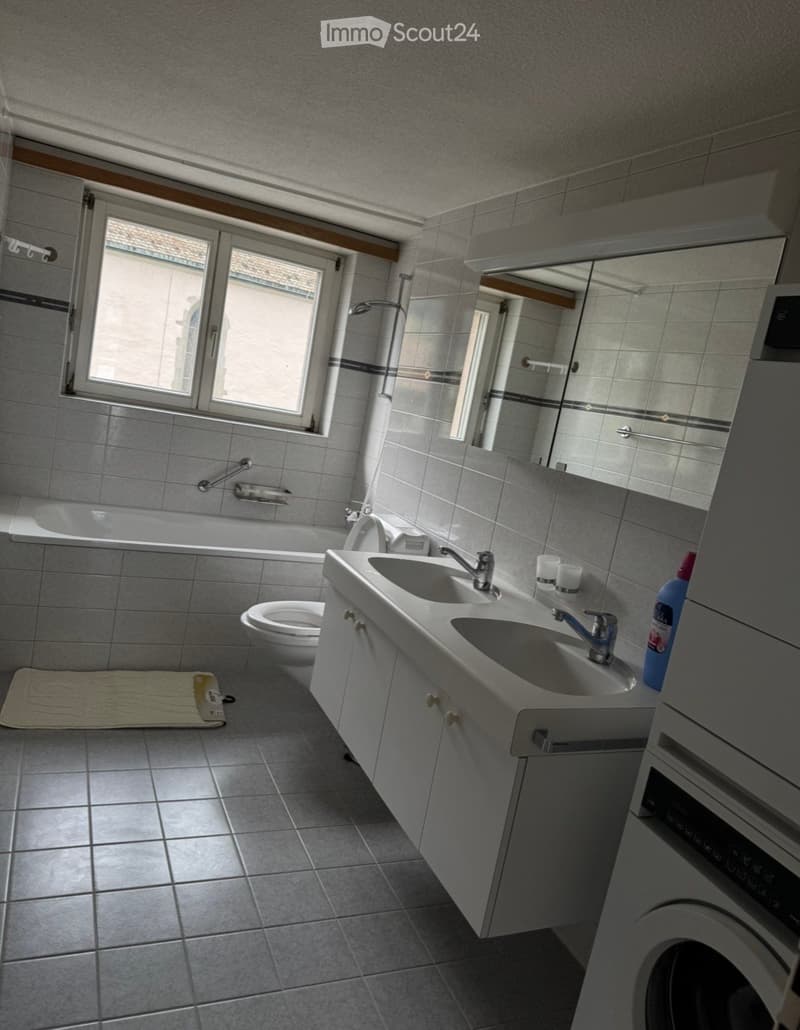 Bathroom with a bathtub, toilet, sink, mirror, and a washing machine. The room features tiled walls and floor, a window, and a towel on the floor.
