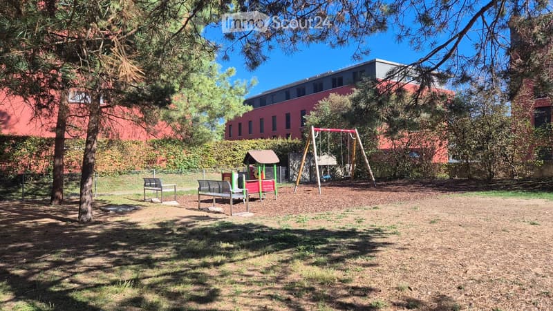 Outdoor playground area with swings, a small playhouse, benches, and trees, surrounded by a red brick building.