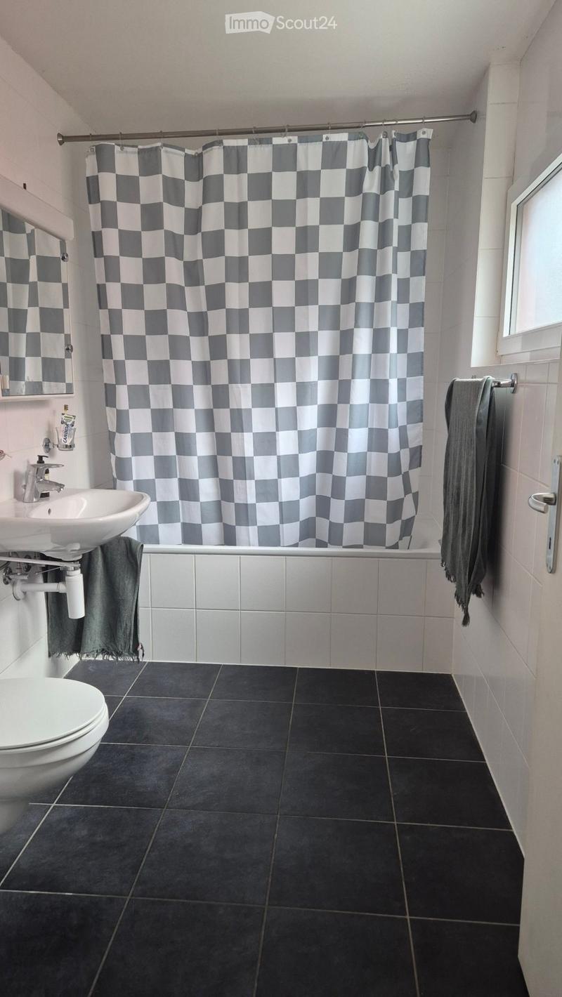 Modern bathroom with black tiled floor, white sink, toilet, and grey shower curtain. Towels are placed on both sides of the sink and bathtub.