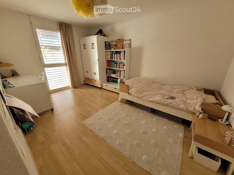 Cozy bedroom with wooden flooring, a single bed with bedding, a nightstand with a lamp, a bookshelf, a wardrobe, and a soft rug.
