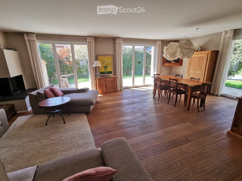 Open-plan living and dining area with wooden flooring, large windows, and sliding glass doors leading to a garden. Features a grey sectional sofa, coffee table, dining table with chairs, and a wooden cabinet with a lamp and painting on top.