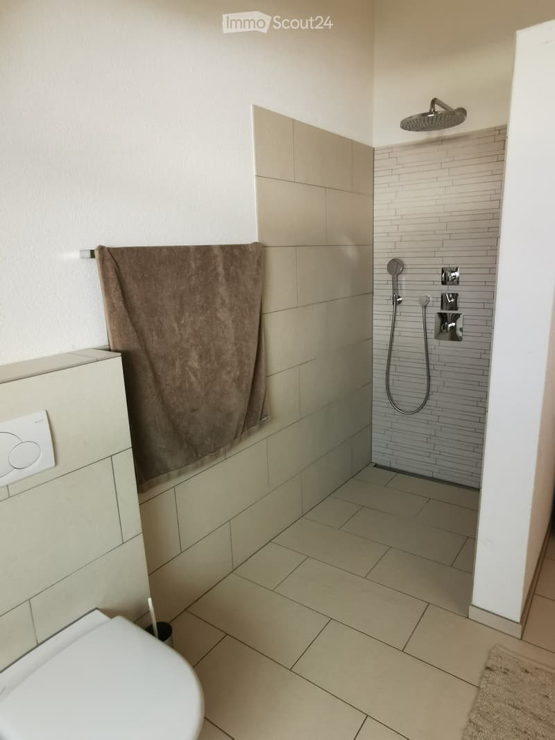 Modern bathroom with beige tiles, a shower area with a handheld showerhead, a toilet, and a towel hanging on the wall.