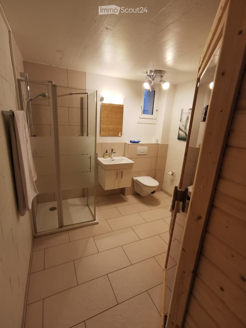 Modern bathroom with tiled floor, glass-enclosed shower, sink with cabinet, toilet, and a wooden-framed mirror. A towel hangs on the left side.