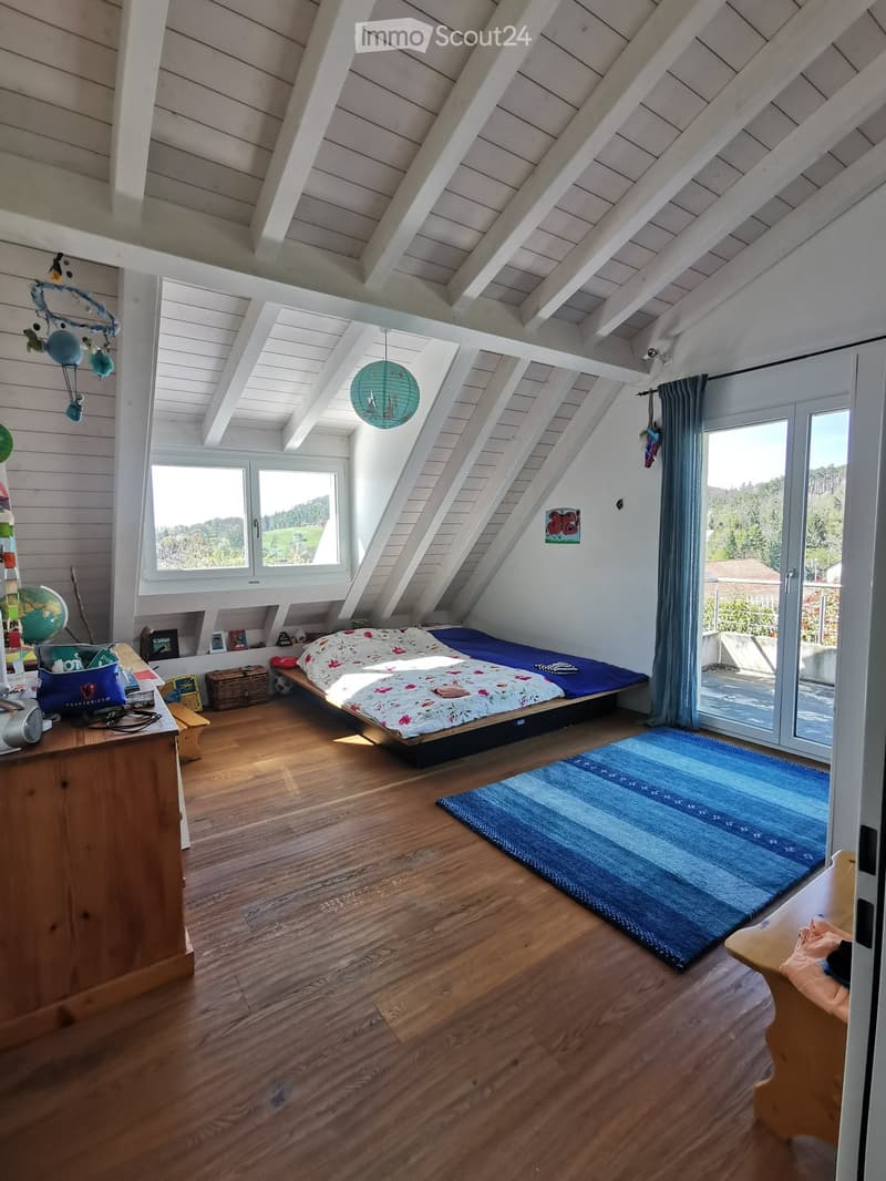 Bright, airy bedroom with wooden floors, slanted white ceiling beams, large windows, and a view of greenery. Features a bed with colorful bedding, a wooden table with various items, a blue rug, and a hanging paper lantern.