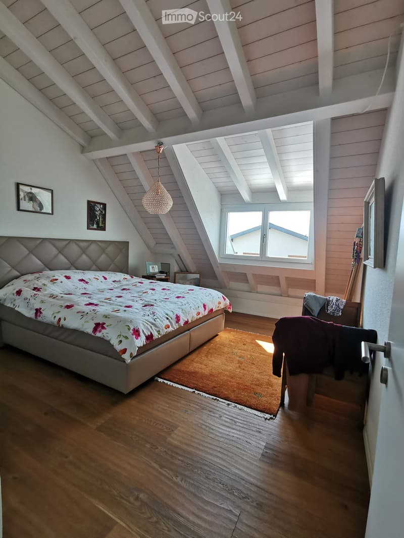 Bright attic bedroom with wooden floors, slanted white ceilings, large windows, a bed with a floral duvet, a nightstand, a mirror, and a pendant light.