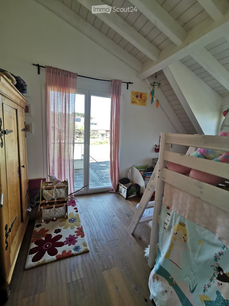 Cozy attic bedroom with wooden floors, slanted ceilings, and large windows. Features a bunk bed with colorful bedding, a wooden wardrobe, a toy box, and a floral rug.