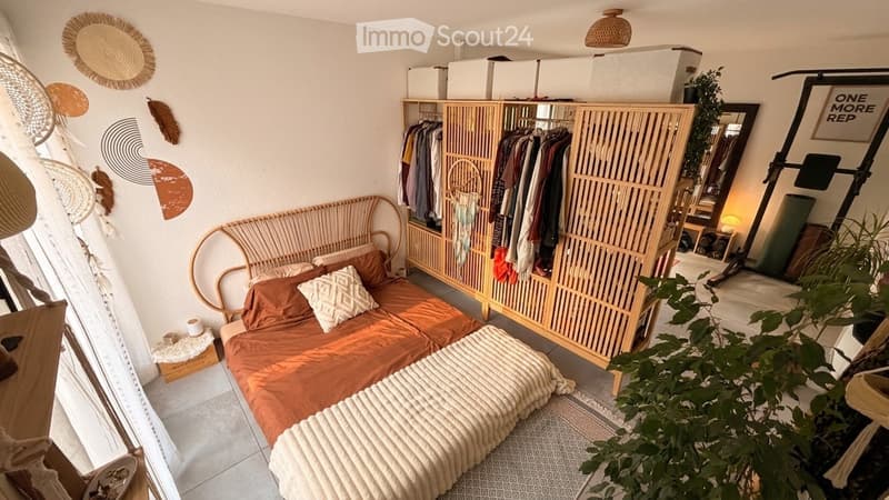 Cozy bedroom with a wicker bed, wooden wardrobe, and decorative wall art. Features a large mirror, plants, and a woven lamp.