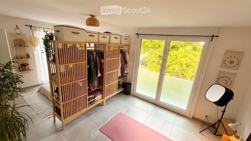 A well-lit room with a large sliding glass door leading to a balcony, featuring a wooden wardrobe, a round mirror on a tripod, a hanging plant, storage boxes, and a small bed area.