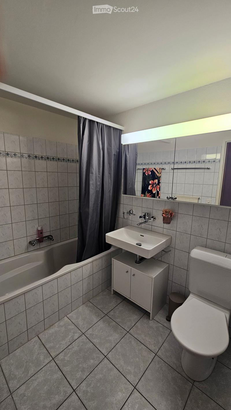 Modern bathroom with a white sink, toilet, bathtub, and mirror. Features tiled walls and floor, a shower curtain, and a small cabinet under the sink.