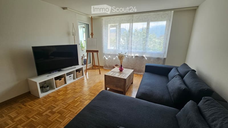 Modern living room with wooden flooring, large windows, a grey sectional sofa, a wooden coffee table with a vase, a flat-screen TV on a white shelf, and an easel near the window.