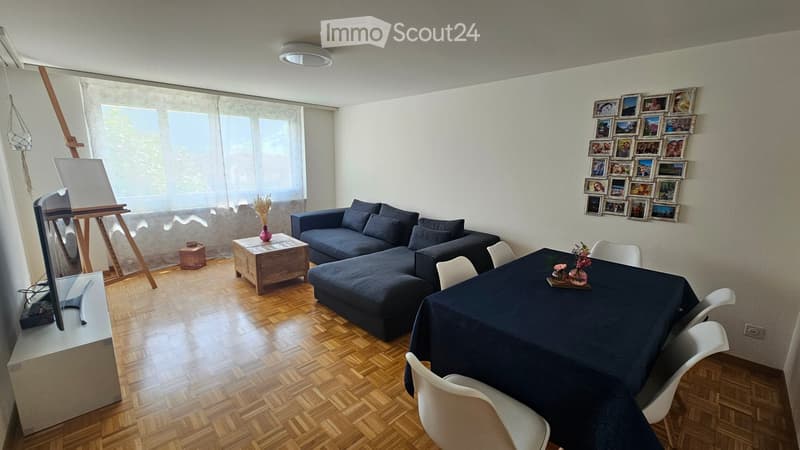 Living room with a dark blue sectional sofa, wooden coffee table, dining table with chairs, TV stand, and an easel with a canvas. The room features wooden flooring and large windows with sheer curtains.