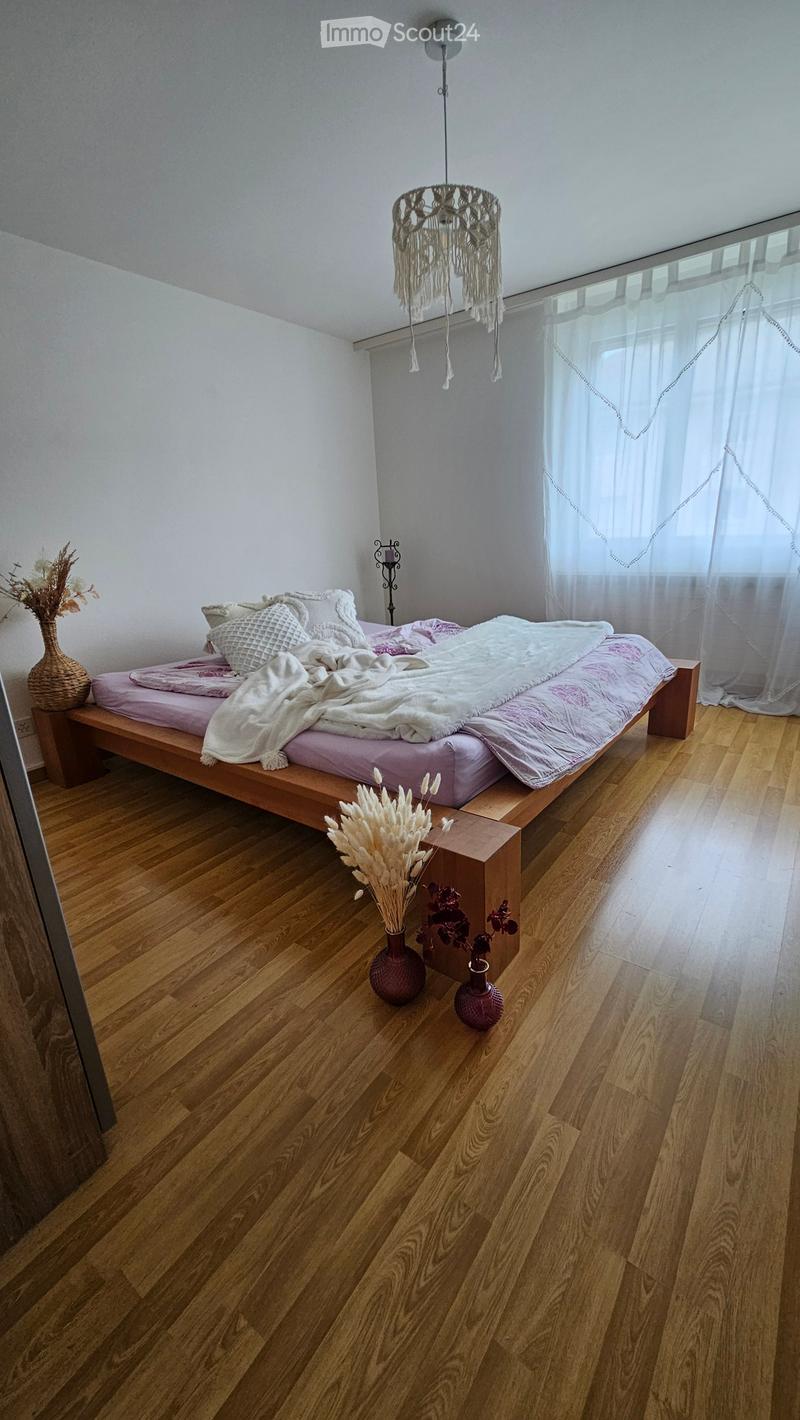 A modern bedroom with a wooden bed, pink bedding, and decorative vases on the floor. The room features wooden flooring, white walls, and a hanging chandelier with fringe details.