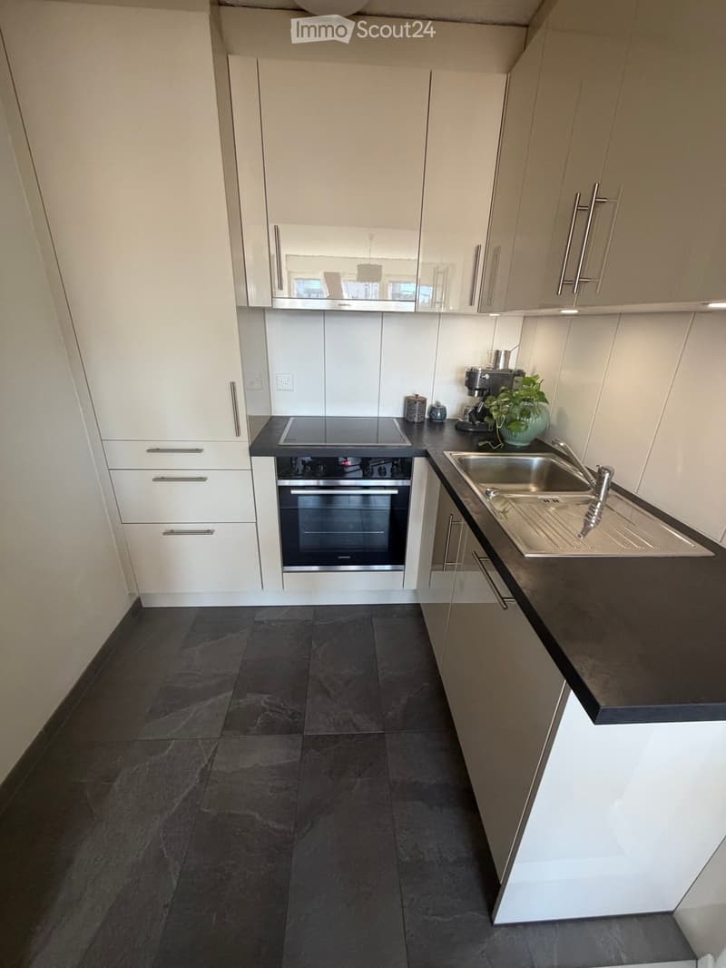 Modern kitchen with white cabinets, dark countertops, stainless steel sink, oven, and coffee maker. Dark tiled floor.