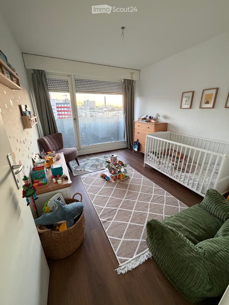 A well-lit children's room with wooden flooring, a green sofa, a crib, a small table with toys, a wicker basket, and a large window with a view of the city.