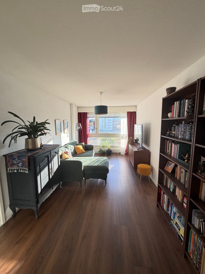 Living room with wooden flooring, a grey sofa with cushions, an ottoman, a bookshelf filled with books, a small yellow stool, a dark cabinet with a potted plant, and a mirror on a side table.