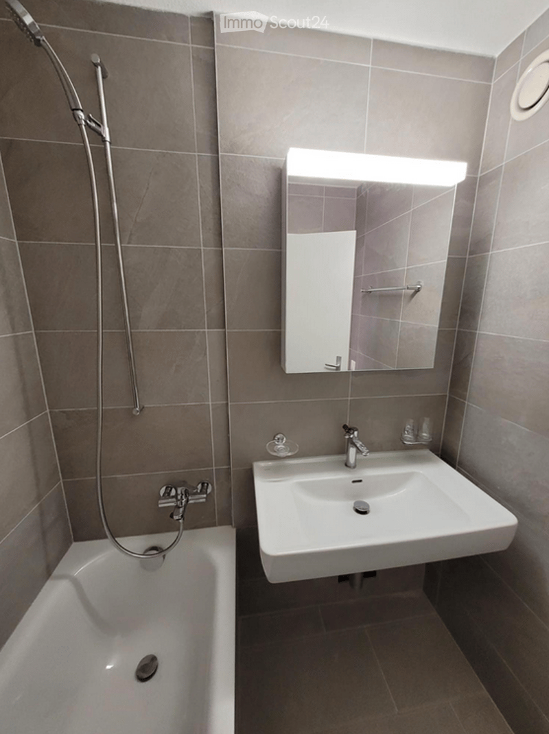 Modern bathroom with a bathtub, sink, mirror, and showerhead. The walls are tiled in a neutral gray color, and the floor is also tiled.