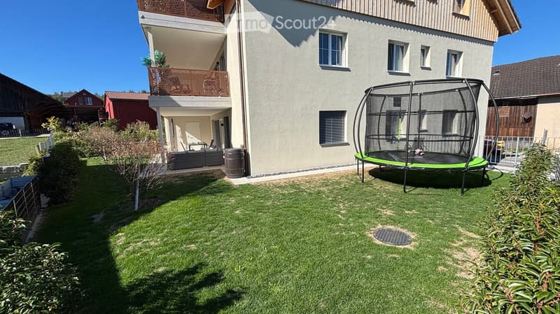 Well-maintained garden with green grass, a trampoline, and a modern house with a balcony and patio seating area.