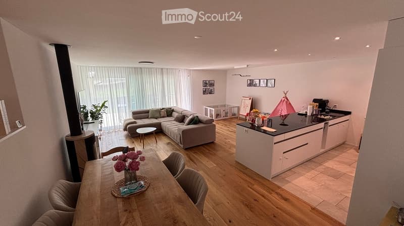 Open-plan living and dining area with a grey sectional sofa, wooden dining table with chairs, kitchen island, and a small playpen. The space features wooden flooring and large windows with sheer curtains.