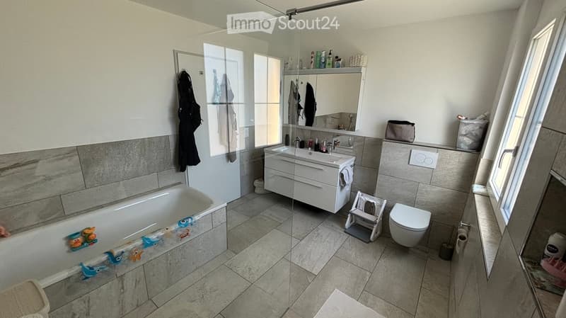 Modern bathroom with a bathtub, sink, toilet, mirror, and a glass shower enclosure. Includes a bathrobe hanging on the wall and a small stool near the toilet.