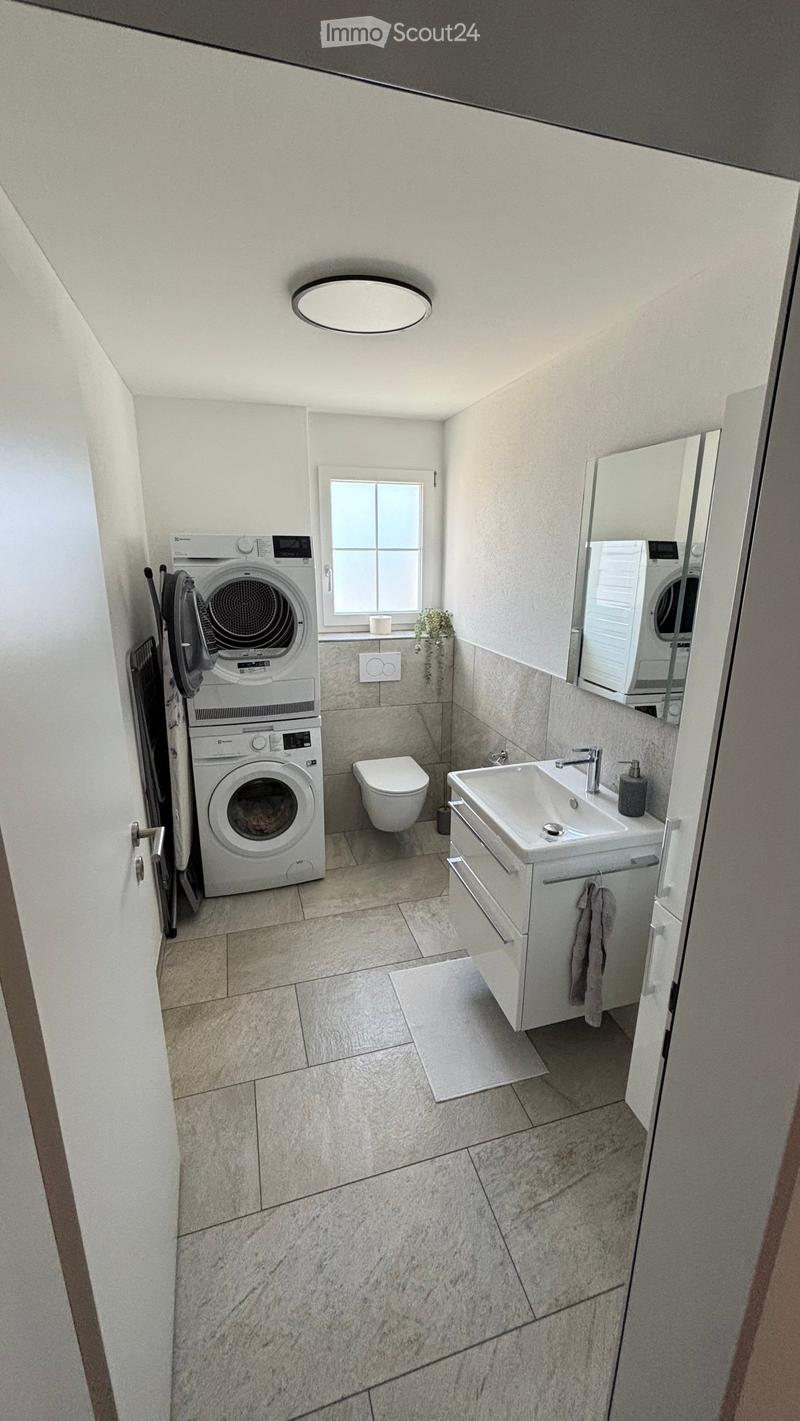 Modern bathroom with a sink, toilet, washing machine, and a mirror. The space features light-colored tiles and a window for natural light.