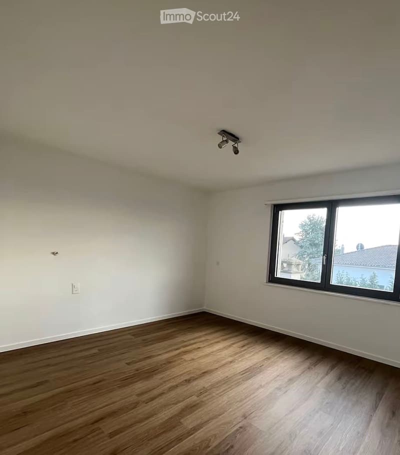 Empty room with wooden flooring, white walls, and a large window showing a view of trees and a neighboring house.