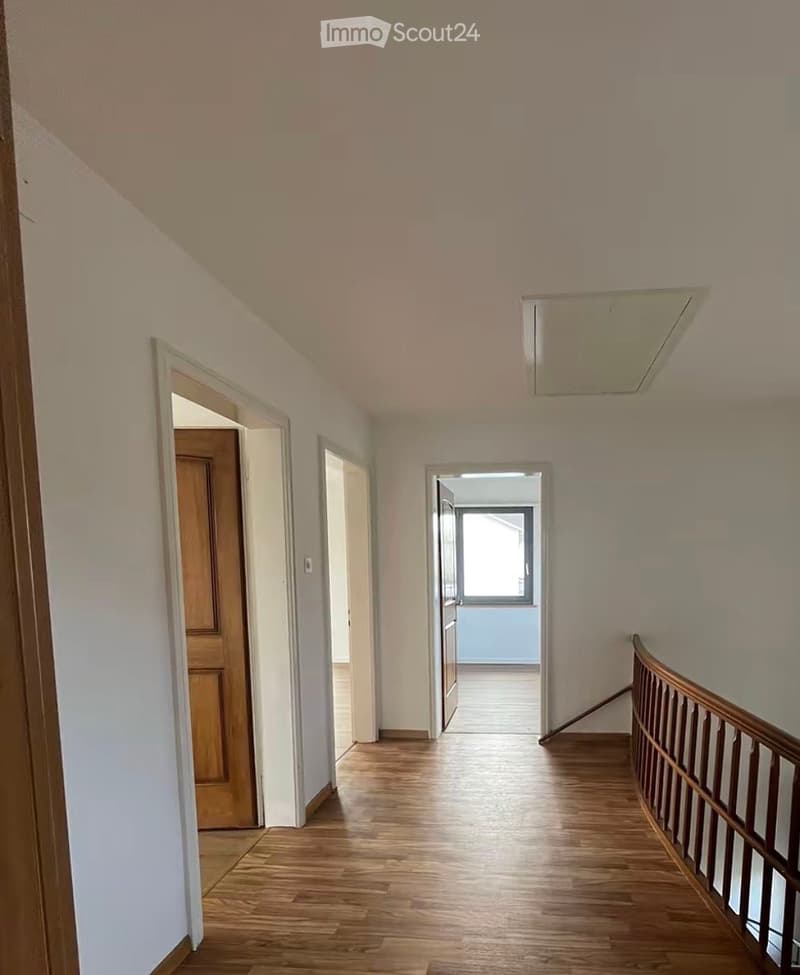 A clean, well-lit hallway with wooden flooring, white walls, and a wooden railing leading to an upper level. There are several doors and a window at the end of the hallway.