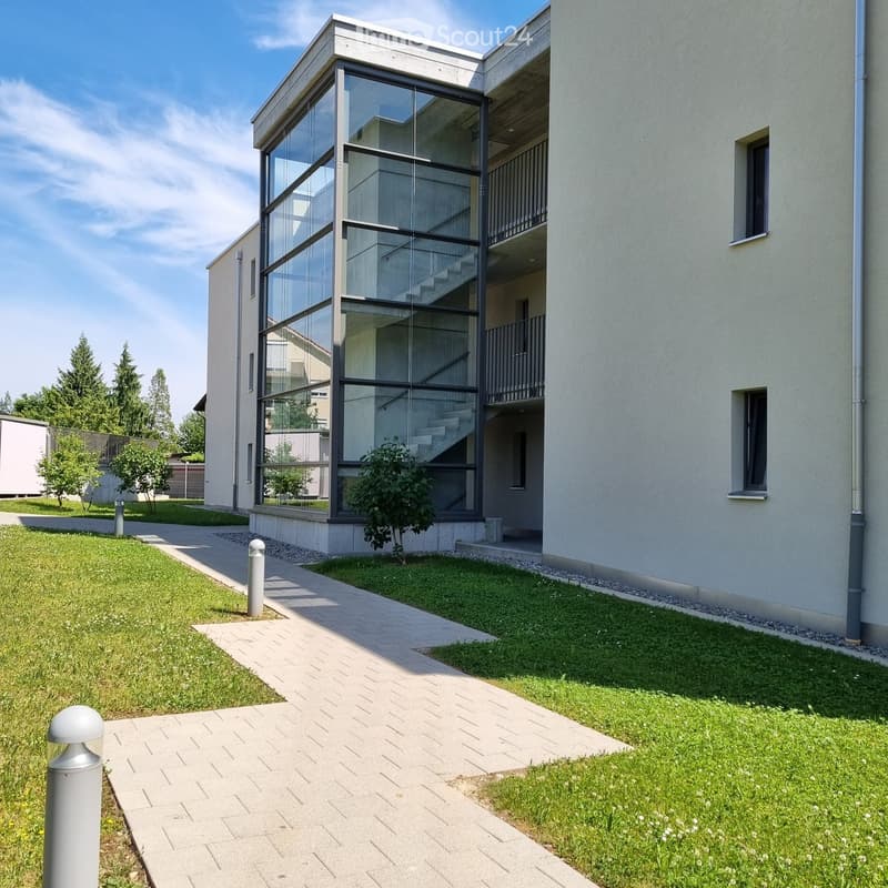 Modern two-story house with a glass staircase leading to an upper balcony, surrounded by a well-maintained lawn and pathway.