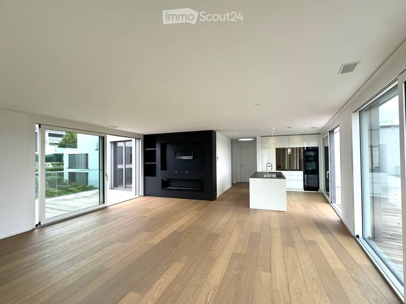 Open-plan living and dining area with light wooden flooring, large glass sliding doors leading to a balcony, a modern kitchen island with a sink, and a built-in fireplace unit.