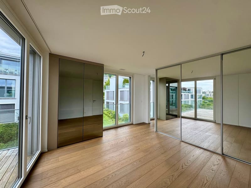 Bright, empty room with wooden flooring, large windows, and sliding glass doors leading to a balcony. The room features a tall, mirrored wardrobe unit and a sleek, modern design.