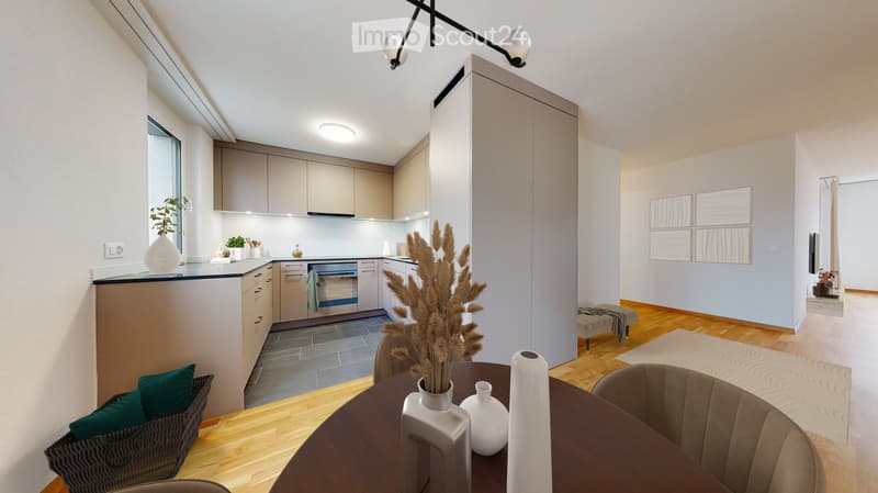 Modern open-plan living and dining area with a round wooden dining table, chairs, decorative vases, a wicker basket, a small kitchen area with cabinets and an oven, and a small seating area with a stool and rug.