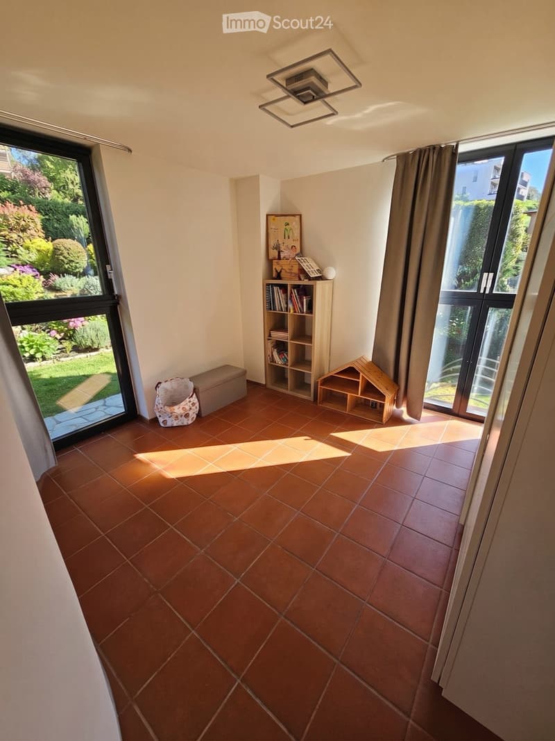 Bright room with terracotta tiled floor, large windows with curtains, a small wooden bookshelf, a toy house, a basket, and a stool.