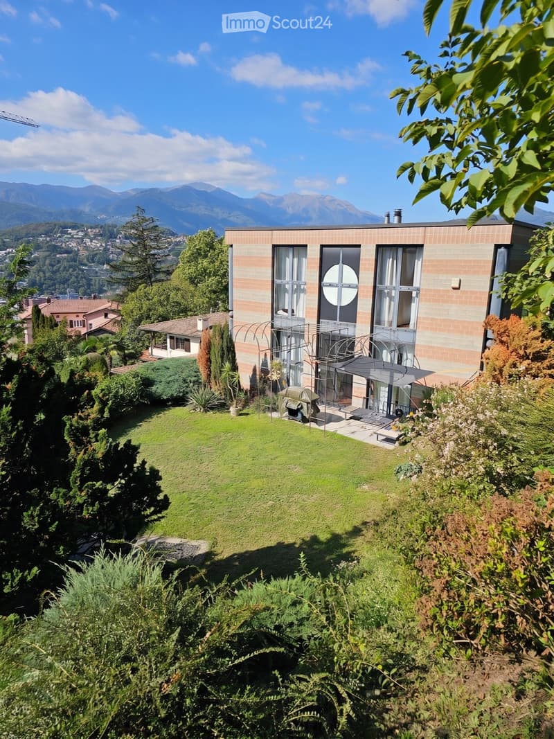 Modern two-story house with brick facade, large windows, and a spacious lawn surrounded by trees and shrubs, overlooking a scenic mountain view.