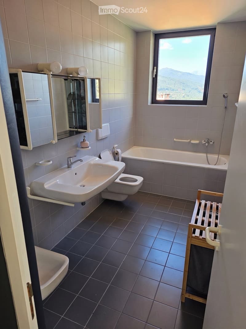 Modern bathroom with a large window offering a scenic mountain view. Features include a white sink, toilet, bathtub, mirror, and a wooden bench.