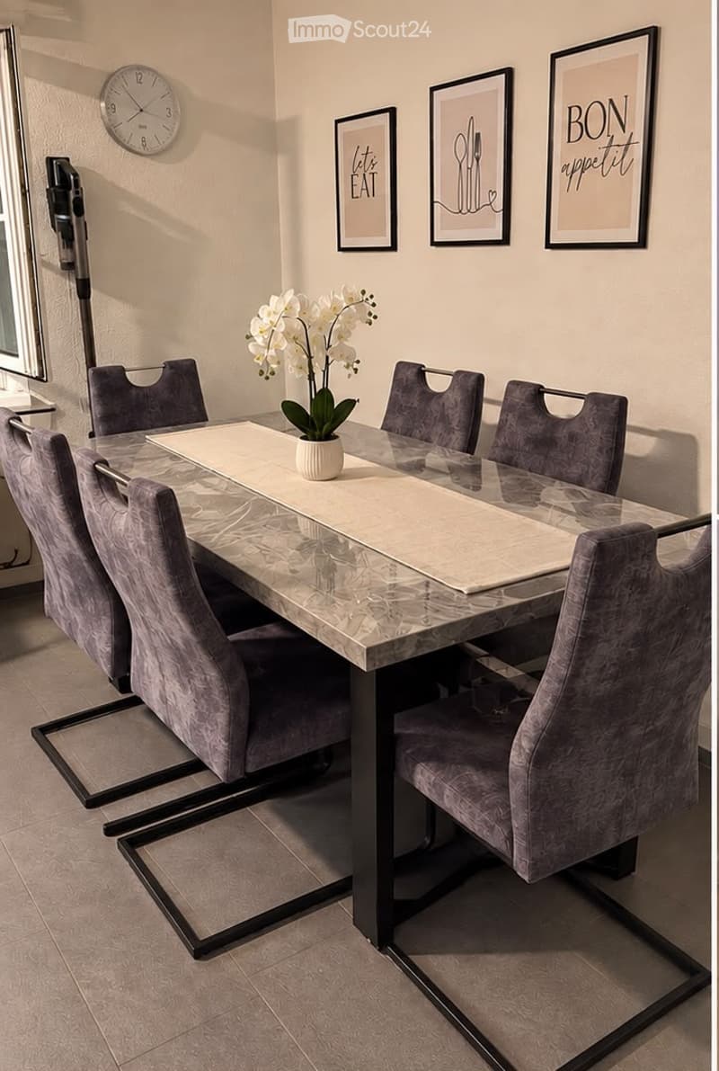 Dining room with a marble table, six grey upholstered chairs, a white flower vase, a wall clock, and three picture frames on the wall.