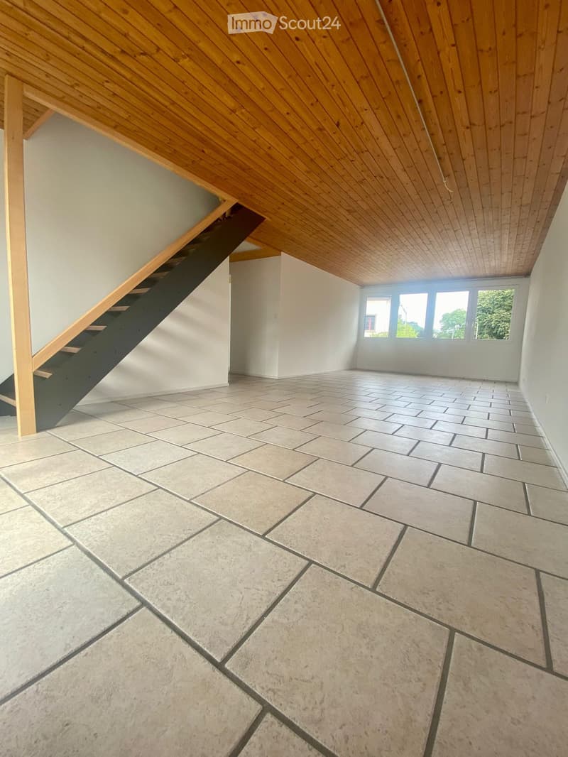 Empty room with tiled floor, wooden ceiling, and a staircase leading upwards. Large windows provide natural light.