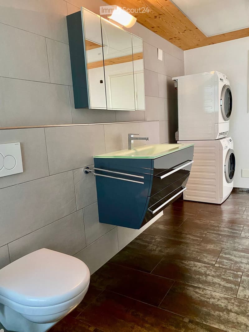 Modern bathroom with a white toilet, glass-fronted sink cabinet, mirrored cabinet, and a stacked washing machine and dryer unit.