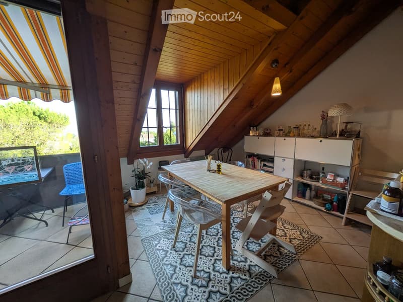 Bright attic room with wooden beams, large window, and skylight. Features a dining table with chairs, a white cabinet, and a patterned rug.