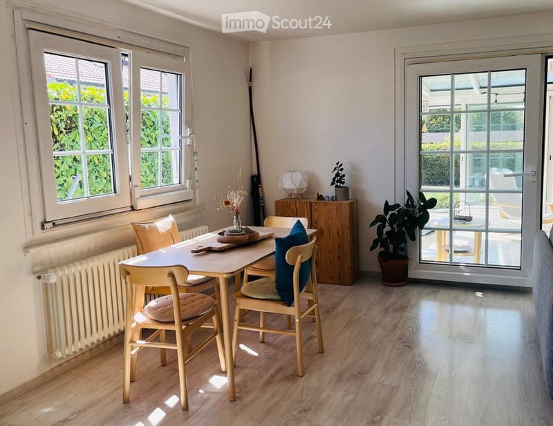 Bright dining area with wooden table, chairs, sideboard, vase with flowers, and potted plant near large windows and sliding glass door leading to outdoor space.