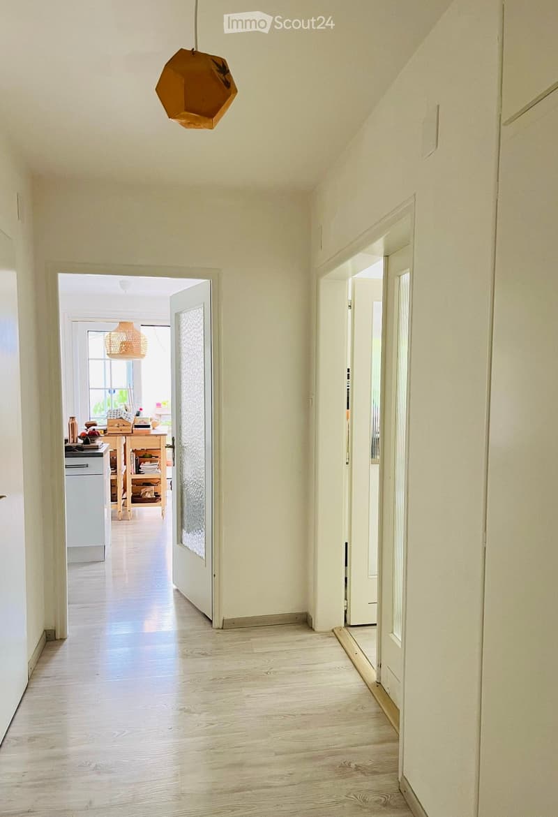 Bright hallway with light wooden flooring, white walls, and a hanging geometric light fixture. Open doors lead to adjacent rooms, including a kitchen area with visible furniture and decor.