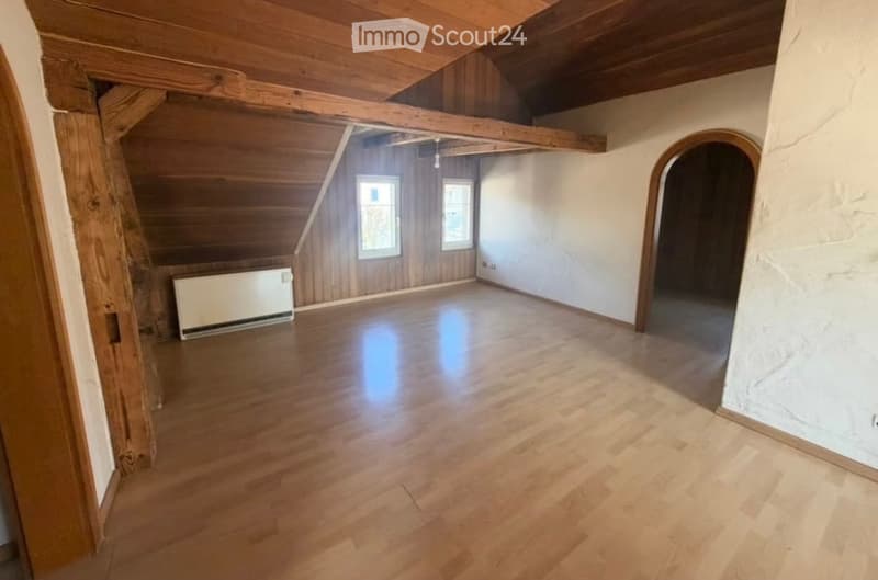 Empty attic room with wooden floors, exposed wooden beams, and two windows. The walls are painted white, and there is a radiator on the left side.