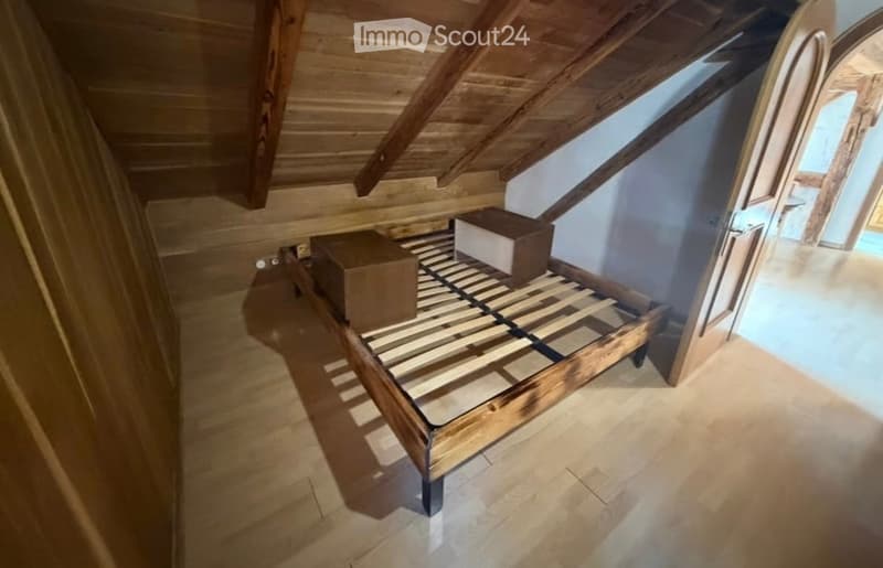 An attic bedroom with wooden slanted ceilings, a wooden bed frame, and a tiled floor. The room is empty except for the bed frame and two boxes placed on it.