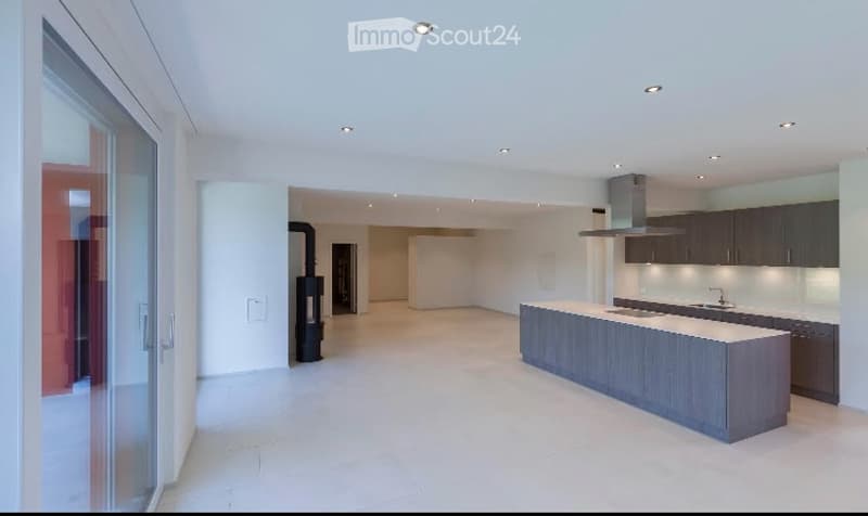 Modern, minimalist kitchen and living area with white walls, light wood flooring, and a large island. The space is completely unfurnished, showing only the structural elements and built-in cabinetry.
